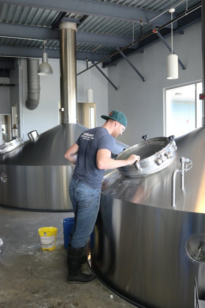 man-wearing-gray-t-shirt-and-blue-jeans-looking-at-stainless-steel-container-1267312 A worker in a brewery inspects large stainless steel fermentation tanks.